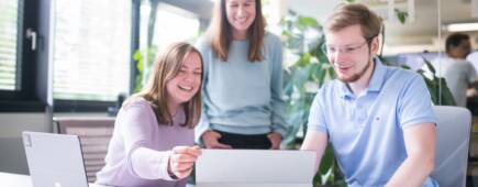 Zwei IT-Expertinnen sitzen mit einem Cloud Architect vor einem Tablet. Sie schauen fröhlich auf den Bildschirm. Neben ihnen steht ein Microsoft Laptop.