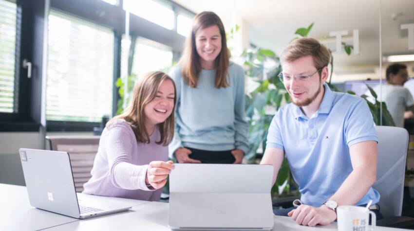 Zwei IT-Expertinnen sitzen mit einem Cloud Architect vor einem Tablet. Sie schauen fröhlich auf den Bildschirm. Neben ihnen steht ein Microsoft Laptop.