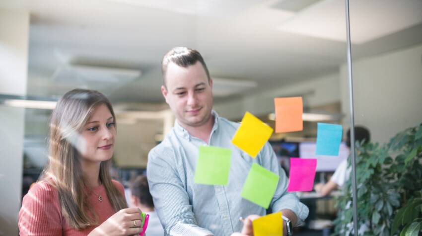 Eine Frau und ein Mann kleben Post-its an die Glasscheibe eines Meetingraums und schauen dabei konzentriert.