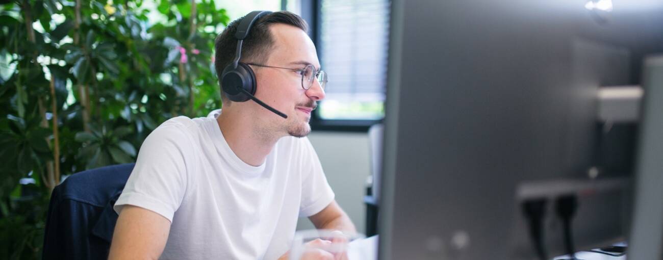 Man sitzt vor einem Bildschirm, hat ein Headset auf und folgt einer Präsentation.